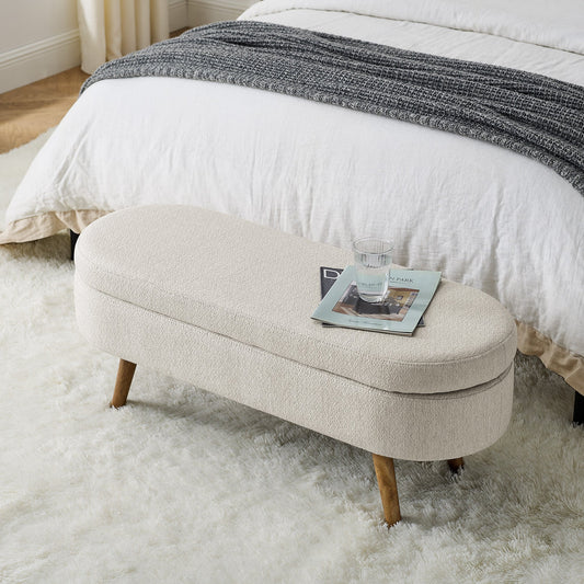 Beige ottoman with a magazine on a fluffy white rug in front of a bed.