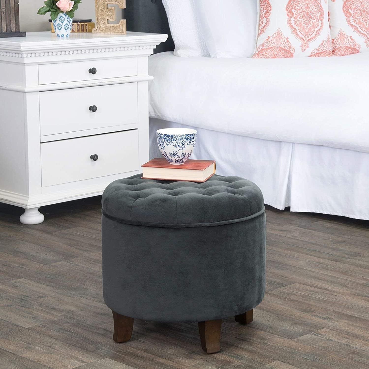 Gray tufted ottoman with a book and cup on top in a bedroom setting.