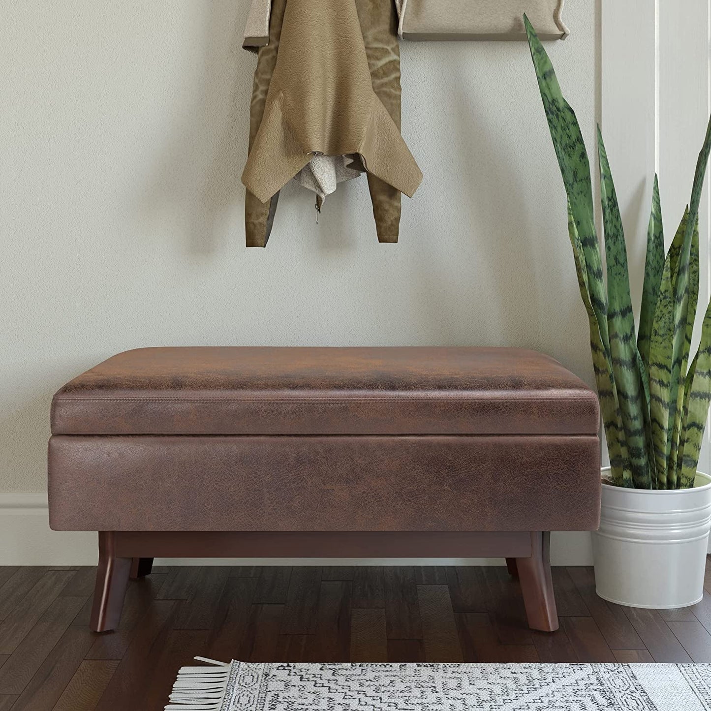 Brown storage ottoman in a room with a plant and coat on a hook.