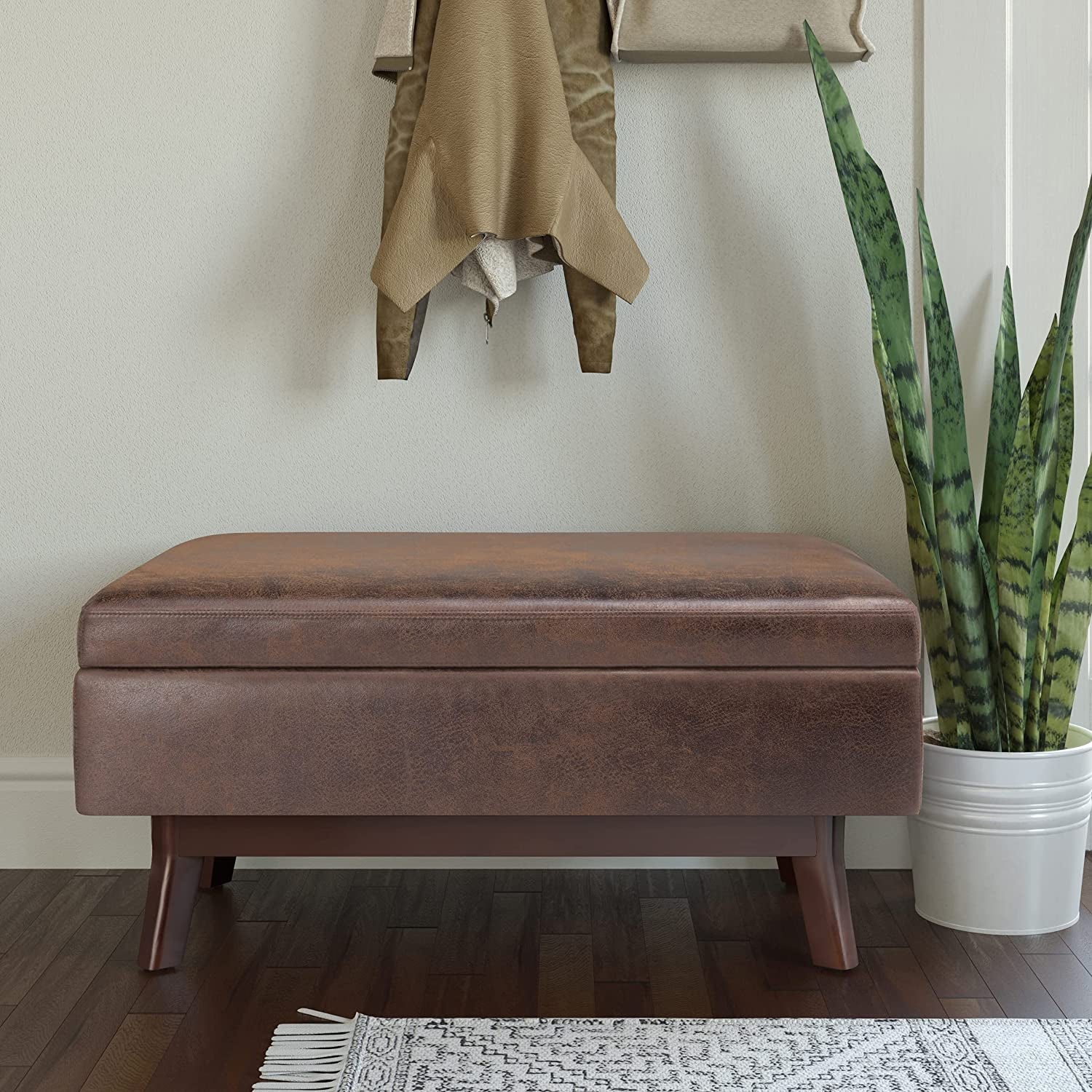 Brown storage ottoman in a room with a plant and coat on a hook.