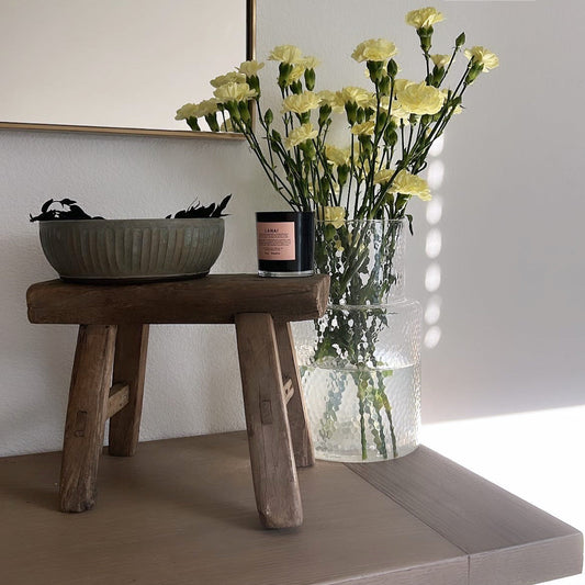 Wooden stool with a vase of yellow flowers and a candle on a light surface.