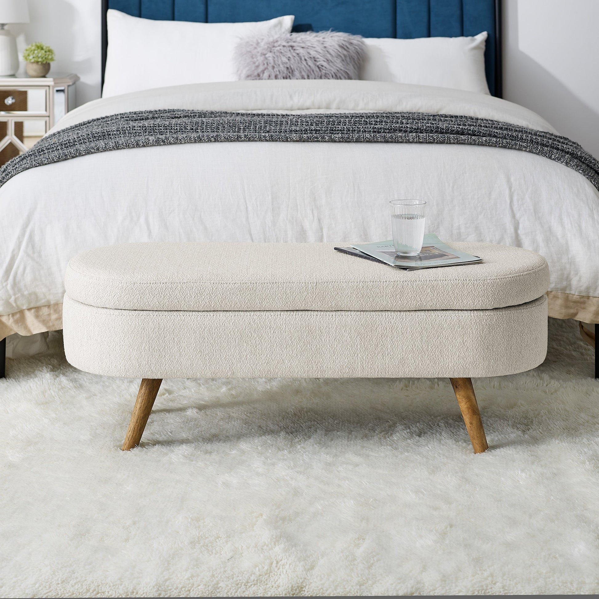 Beige ottoman with wooden legs in front of a bed with white bedding and a blue headboard.