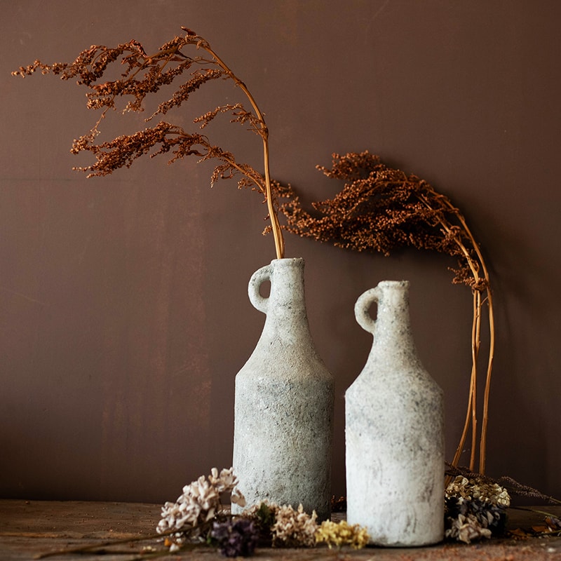 Two stone-vase-like bottles with dried plants against a brown background