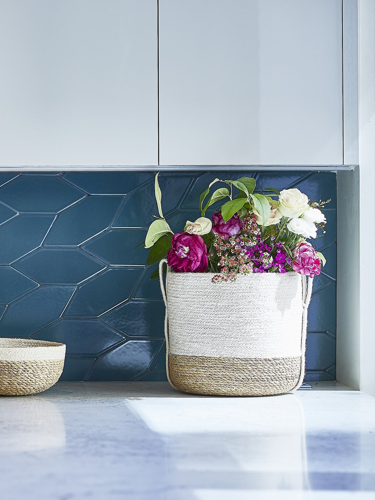 Decorative woven basket with flowers on a kitchen counter against a blue tiled wall.