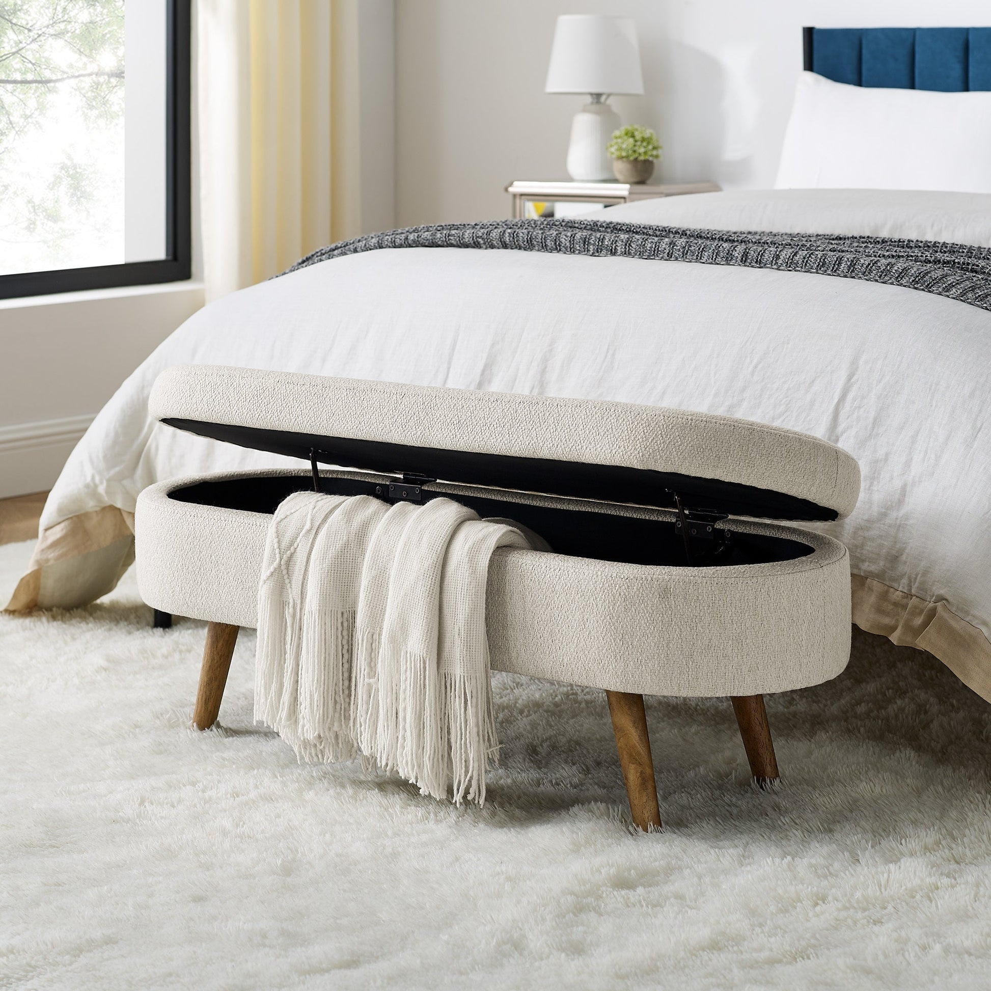 Beige upholstered storage bench with wooden legs in a bedroom setting.