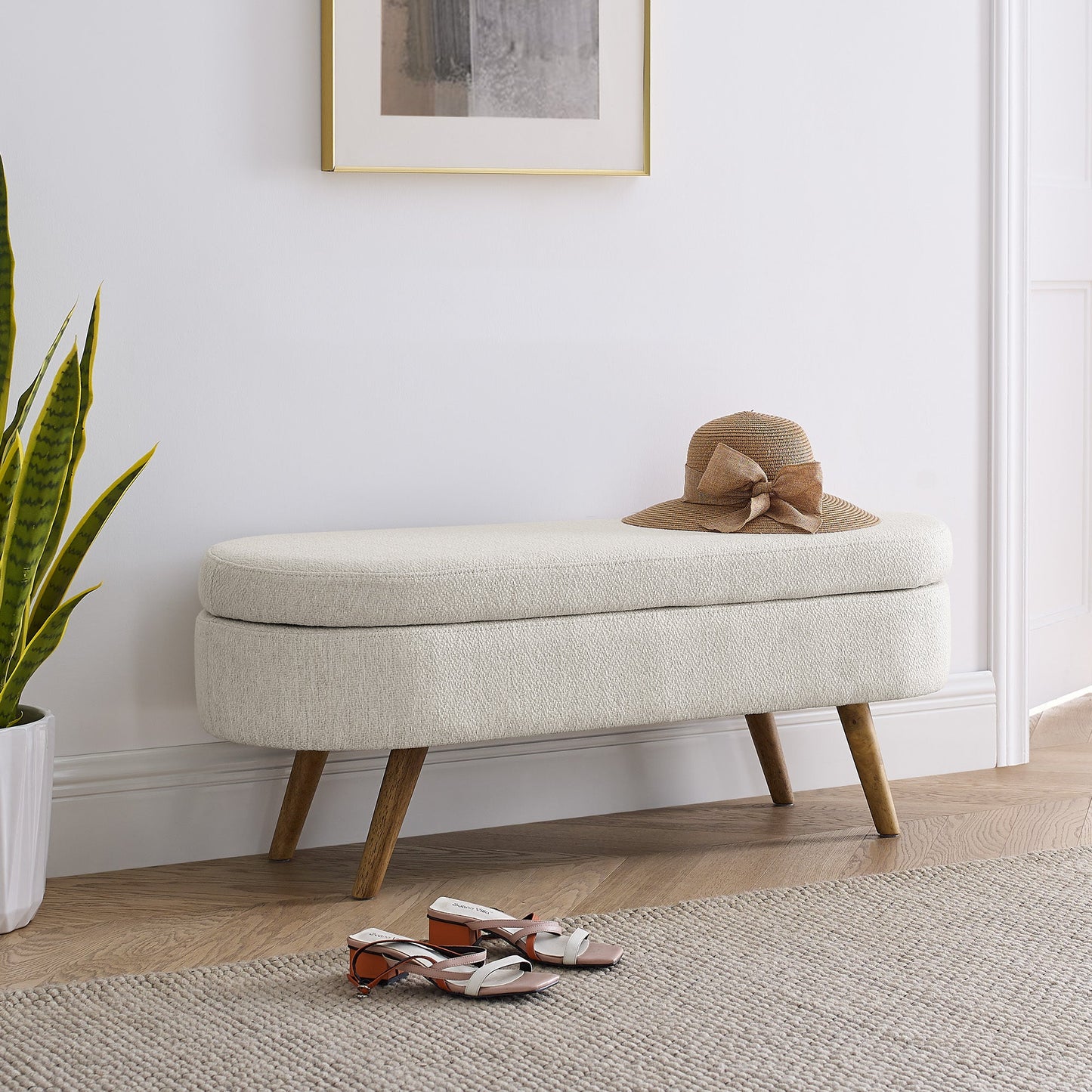 Beige storage bench with a hat on top in a room with a plant and shoes on the floor.