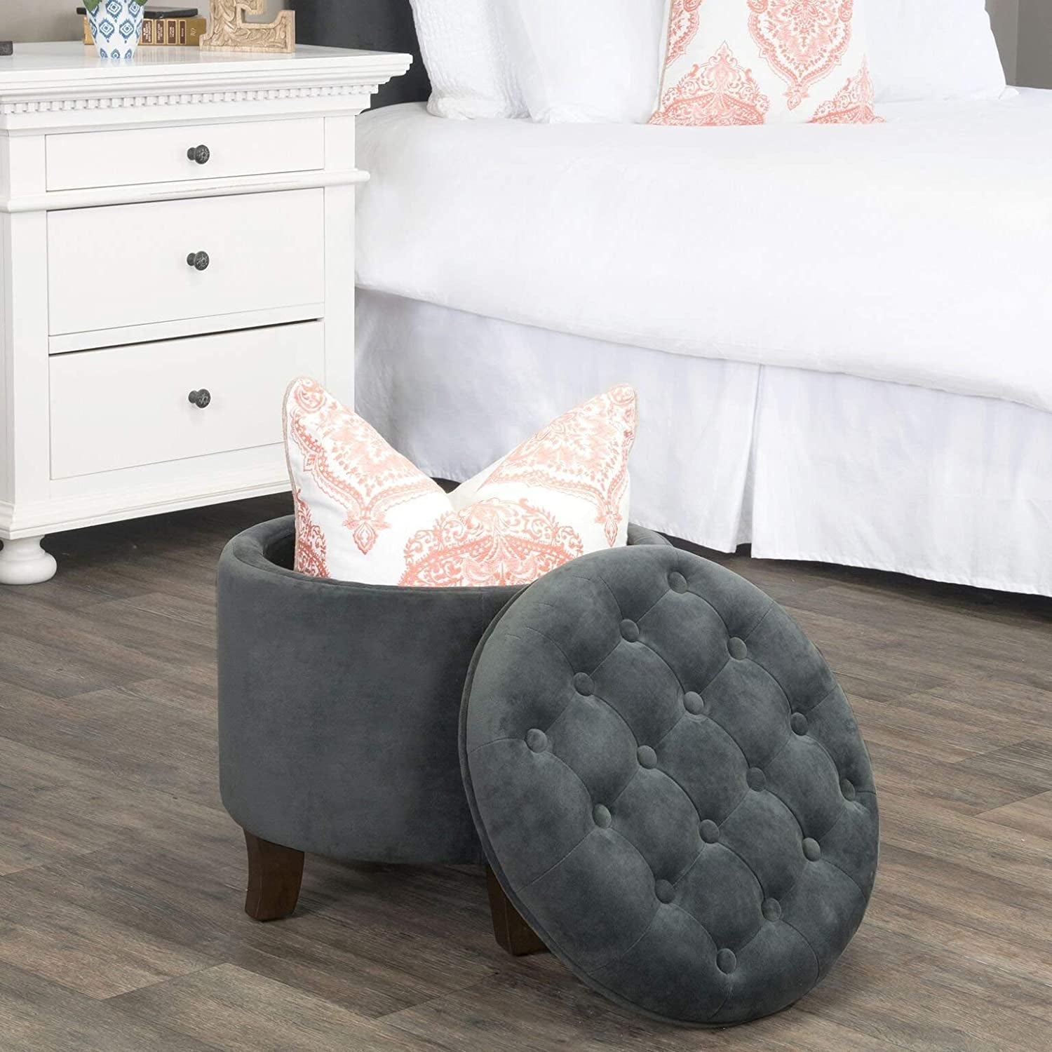 Gray tufted ottoman with a lid, containing decorative pillows, in a bedroom setting.