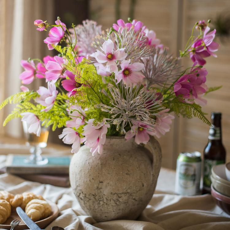 Decorative floral arrangement in a textured vase on a table with food and drinks.
