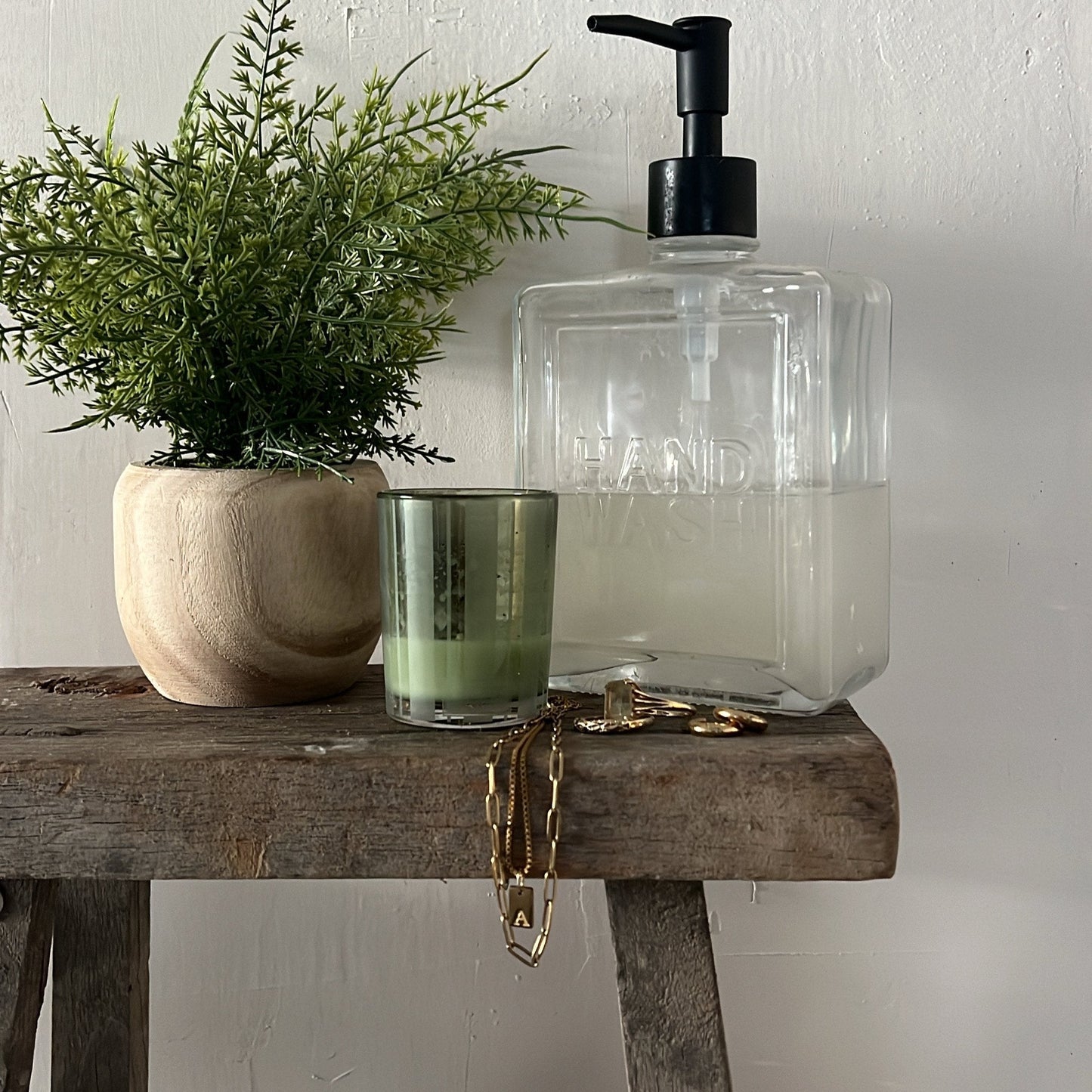 Clear glass soap dispenser with black pump on a wooden surface next to a potted plant and candle.
