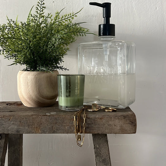 Clear glass soap dispenser with black pump on a wooden surface next to a potted plant and candle.