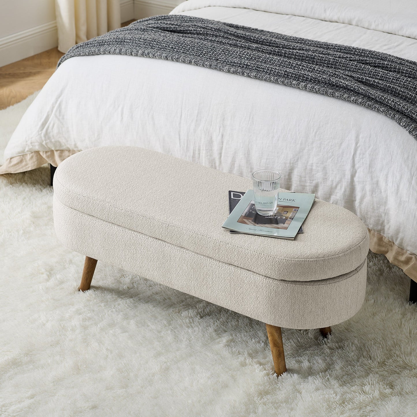 Beige ottoman with a magazine on a fluffy white rug in front of a bed.